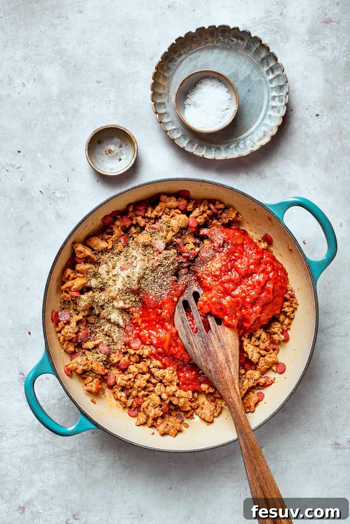 Rich tomato sauce is being stirred into the pan with the cooked sausage and pepperoni, preparing the flavor base.