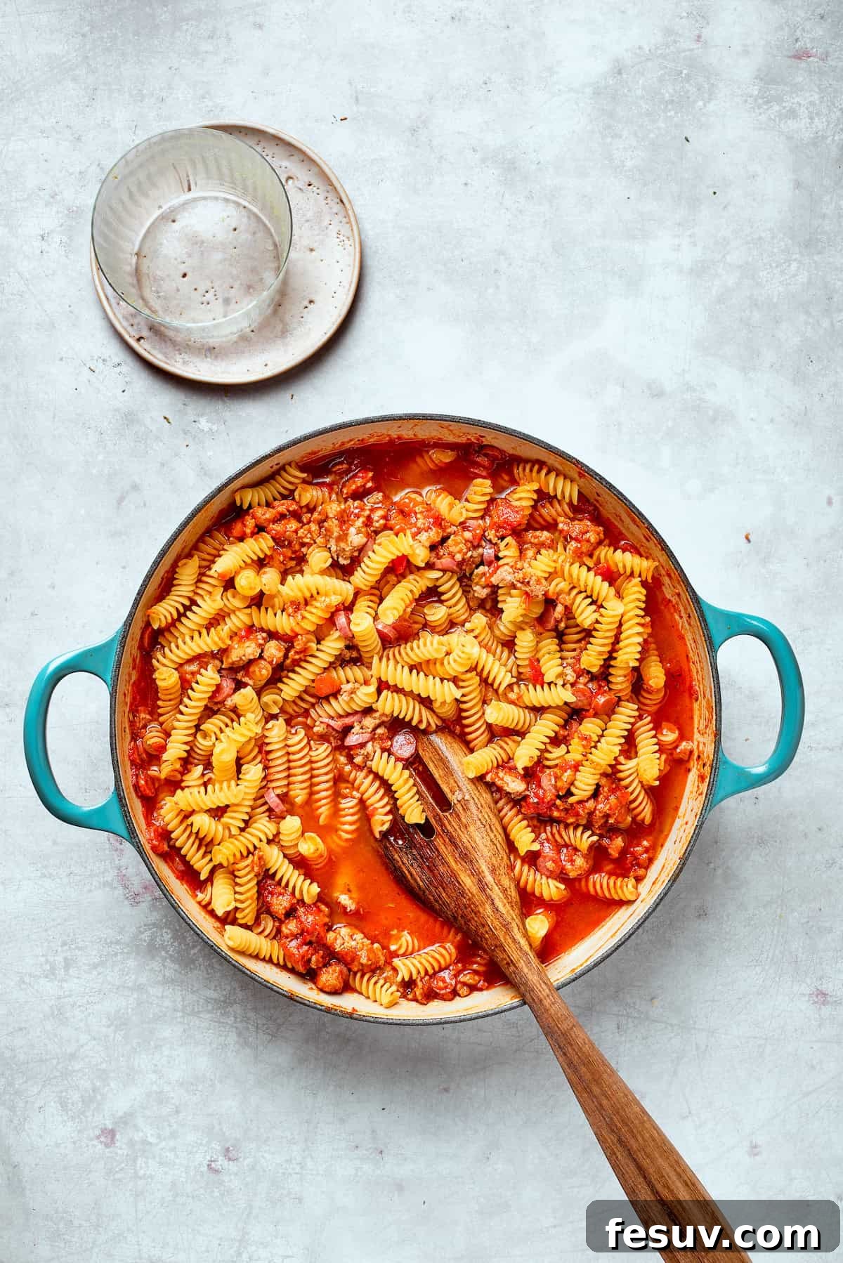 A wooden spoon is used to stir the rotini pasta into the rich tomato and sausage sauce, ensuring it's well coated.