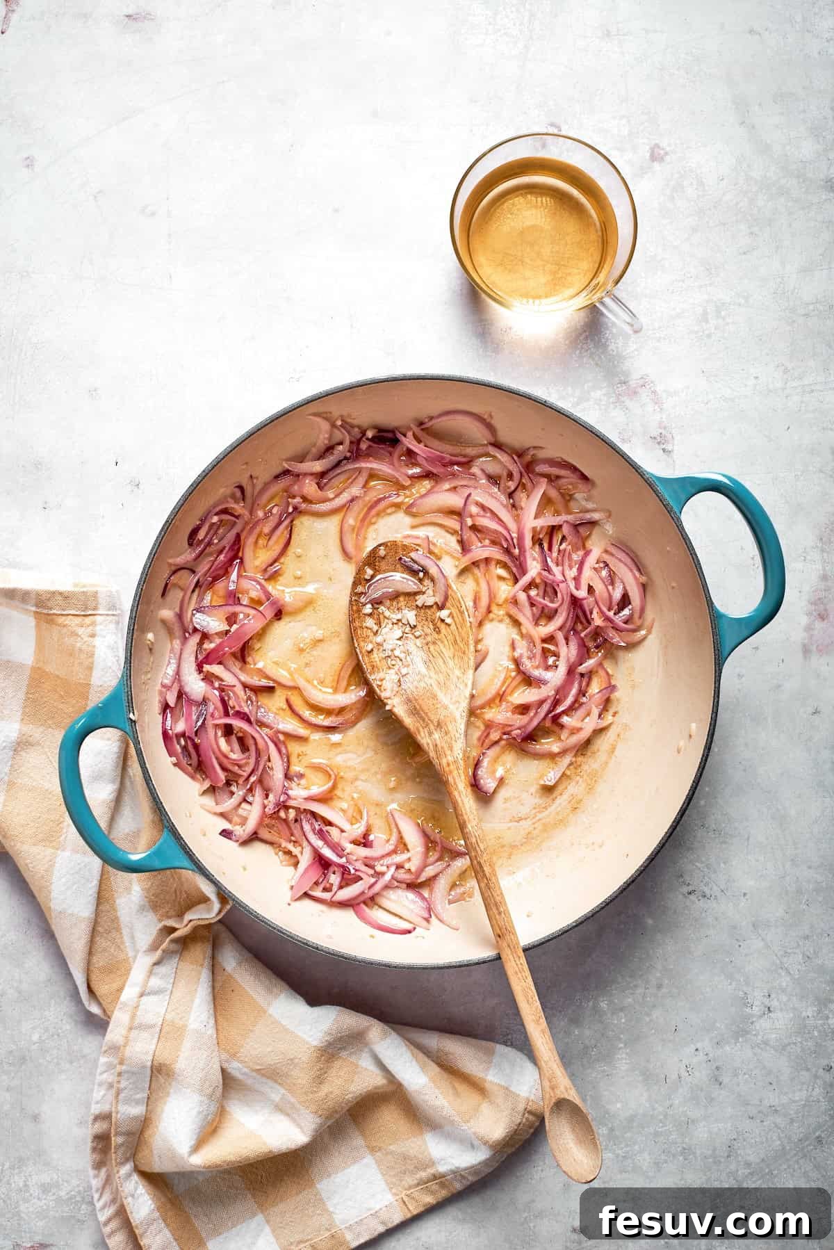 A wooden spoon stirring thinly sliced red onions and minced garlic in a skillet, gently sautéing.