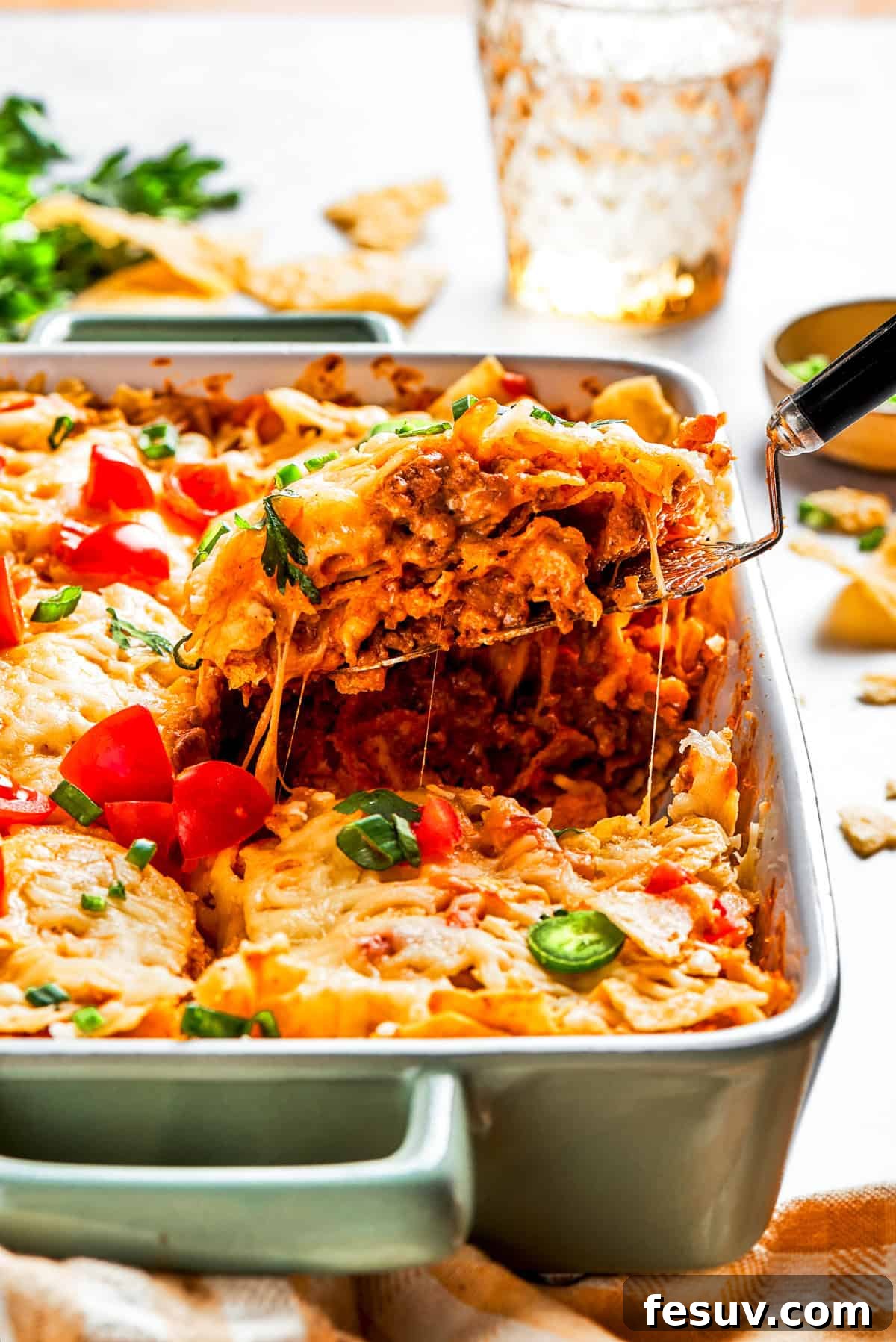 A serving spoon lifting a generous slice of golden-brown Dorito casserole, revealing its cheesy, meaty layers, from a rustic baking dish.
