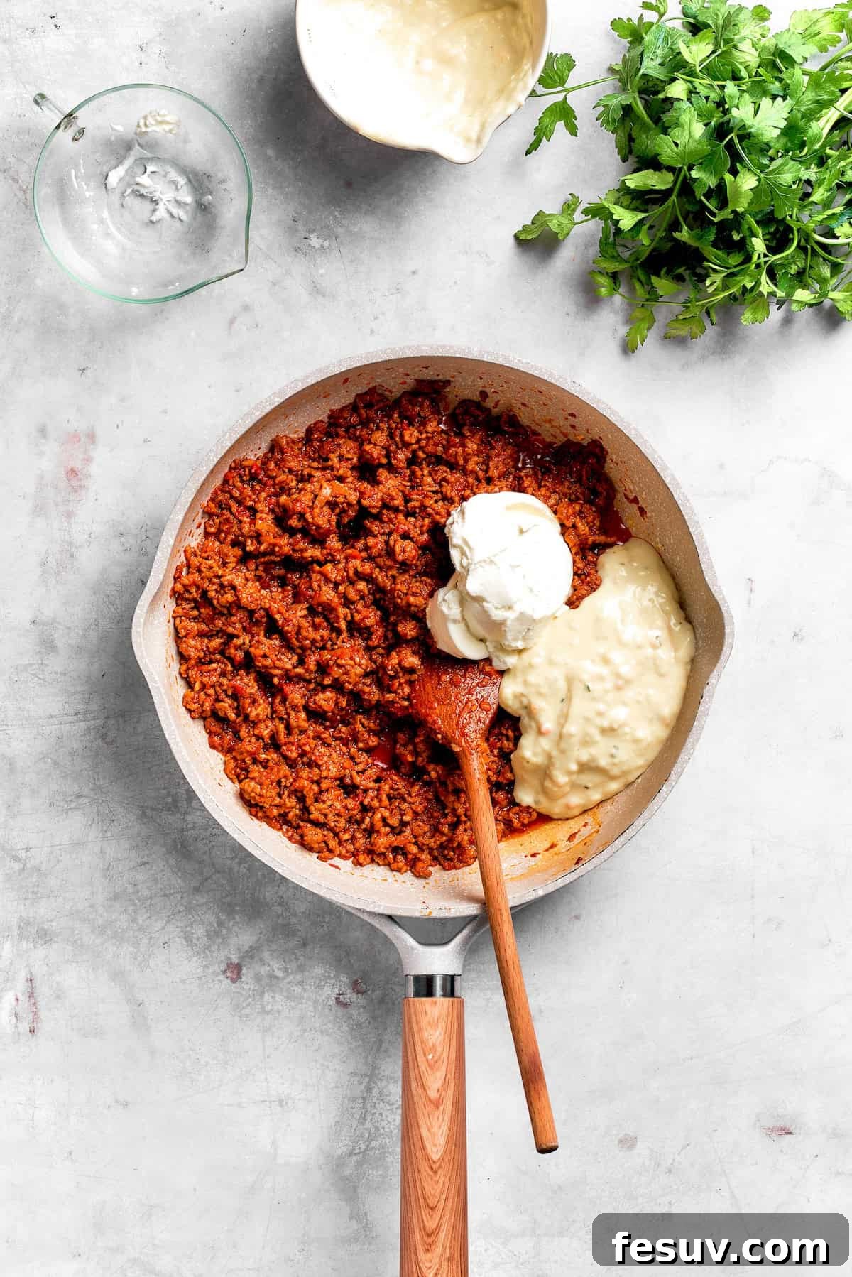 Sour cream and cream of chicken soup being stirred into the seasoned ground beef mixture in a skillet, creating a creamy casserole filling.