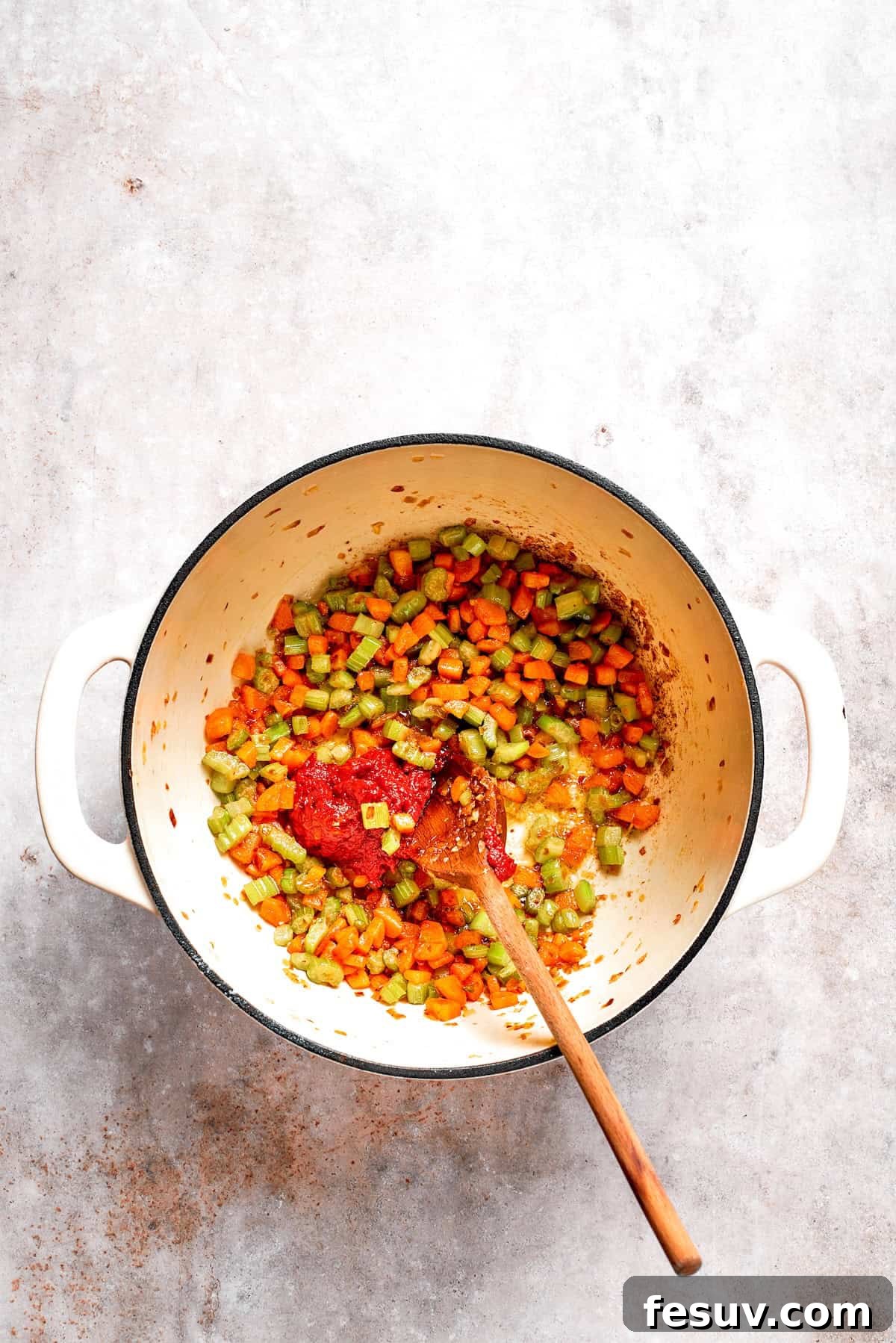 Bright red tomato paste being stirred into sautéed vegetables in a pot, coating them evenly.