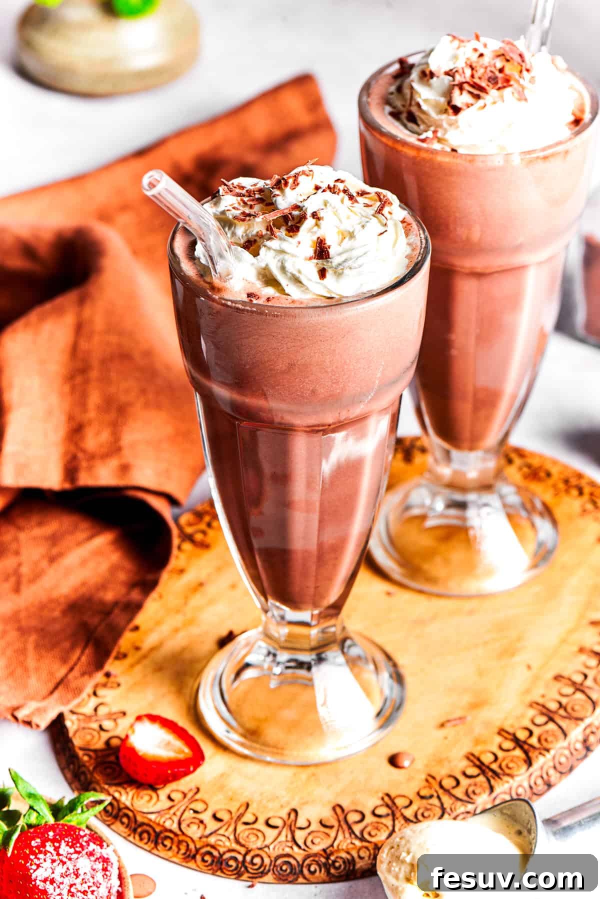 Two exquisitely garnished chocolate milkshakes rest on a rustic wooden tray, adorned with whipped cream, fresh strawberries, and chocolate drizzle, ready to be enjoyed.
