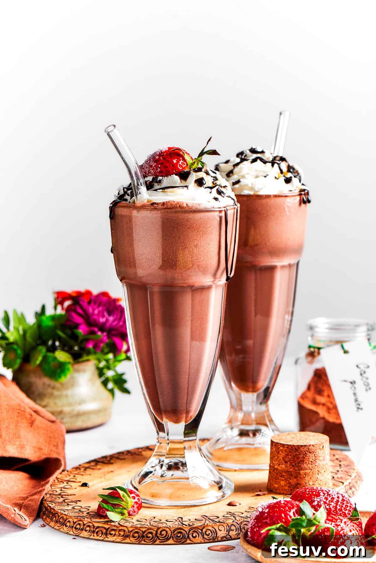 Two perfectly prepared chocolate milkshakes, adorned with generous dollops of whipped cream, fresh strawberries, and a delicate chocolate drizzle, served in tall, frosted glasses on a dark wooden tray.