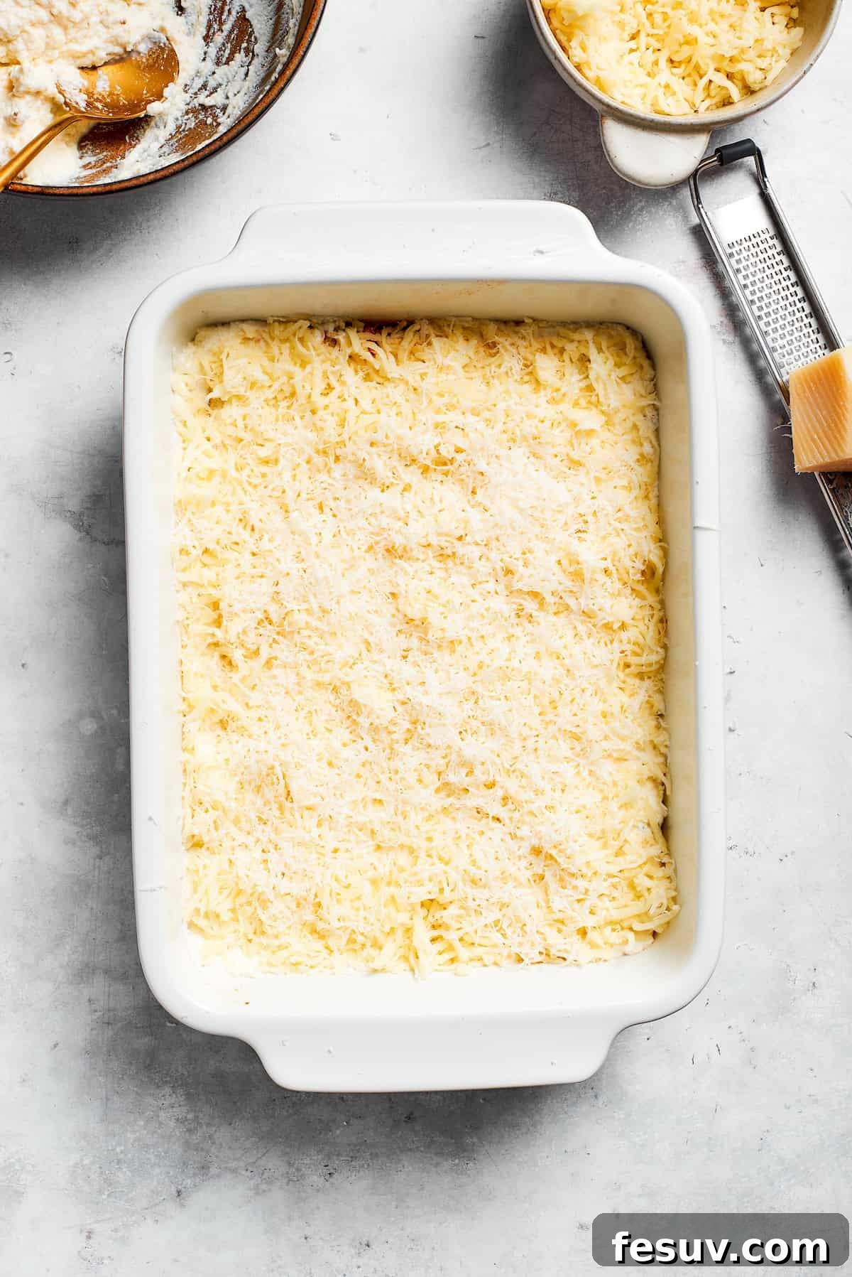 Shredded cheese is added to the ricotta layer of the lasagna, preparing for the next layer.