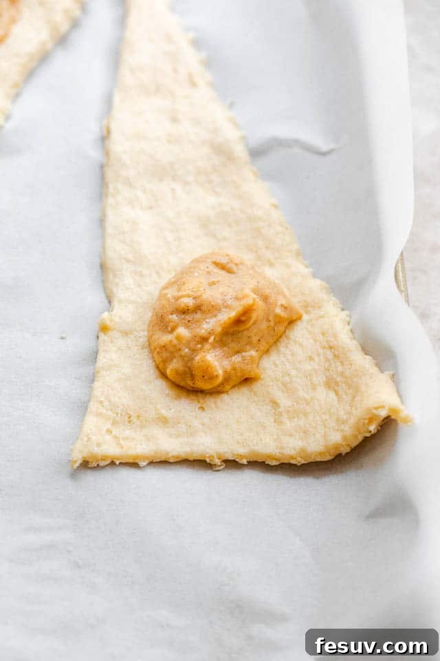 These Pumpkin Crescent Rolls are the best fall treat! Buttery rolls stuffed with a sweet pumpkin cream cheese filling and drizzled with vanilla glaze. Unrolled raw crescent dough with a dollop of pumpkin filling on the wider end, ready to be rolled.