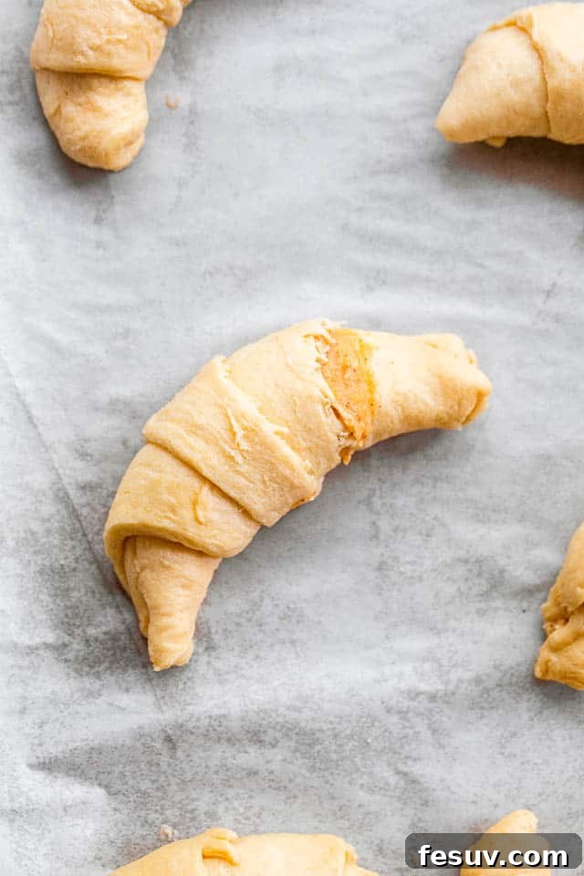 These Pumpkin Crescent Rolls are the best fall treat! Buttery rolls stuffed with a sweet pumpkin cream cheese filling and drizzled with vanilla glaze. Preparing crescent rolls for baking, showing them rolled up and arranged on a baking sheet.