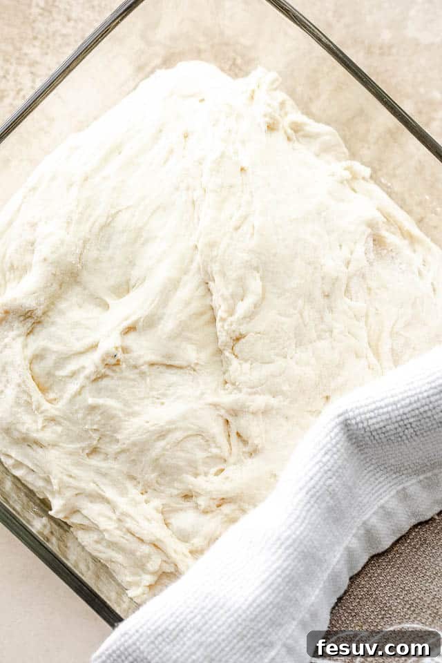 Effortless Artisan Bread 5 No Knead Dough in a clear glass container and halfway covered with a kitchen towel