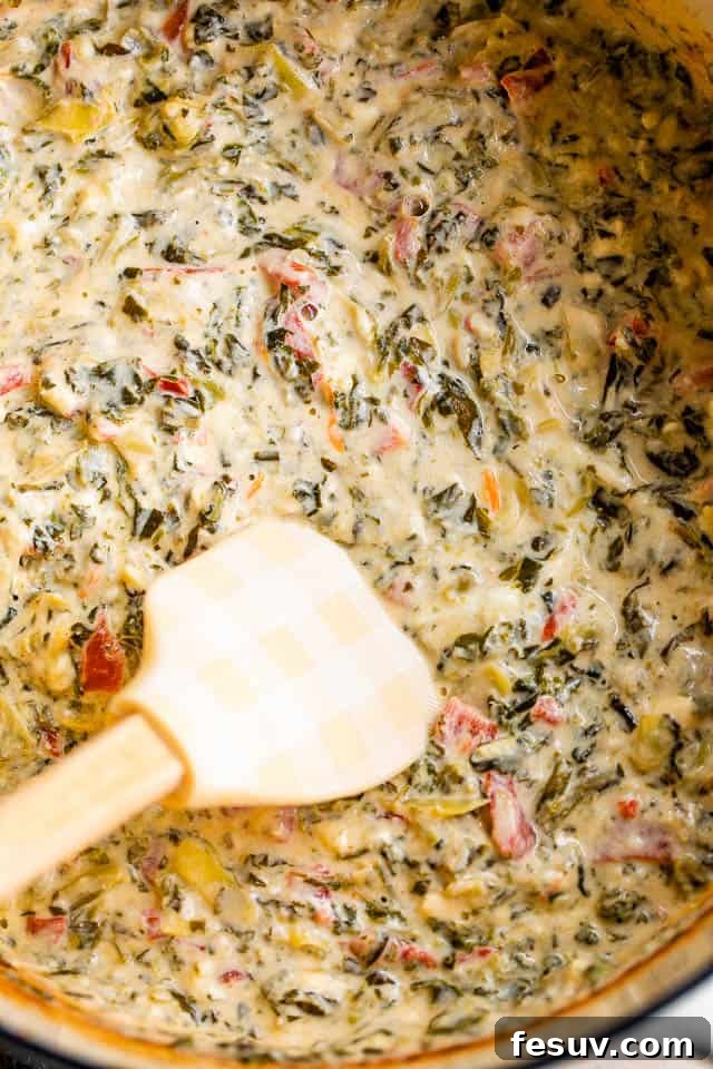 This Slow Cooker Spinach Artichoke Dip is a festive and delicious dip featuring artichokes, sweet red peppers and a medley of cheeses. A spatula stirring creamy spinach artichoke dip in a slow cooker, highlighting the rich texture and melded ingredients.