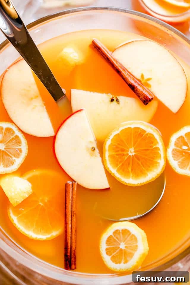 Overhead shot of a full punch bowl of slow cooker apple cider punch, with a ladle resting inside and an abundance of colorful fruit slices (apples, oranges) gracefully floating on the surface.