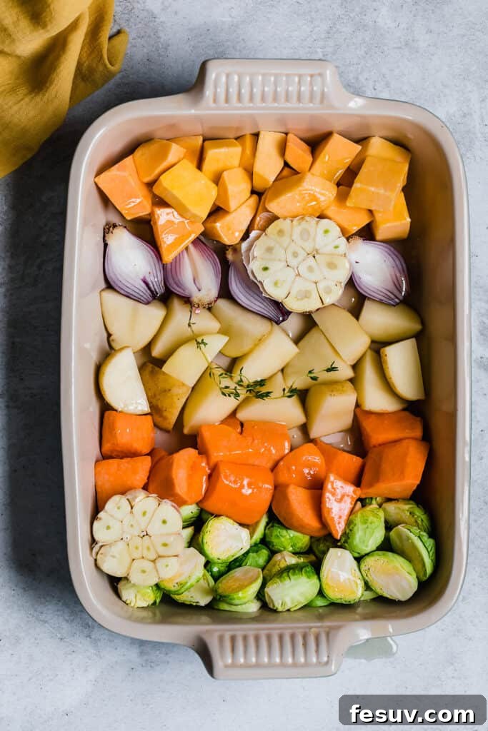 Chopped root vegetables, including butternut squash, potatoes, and red onions, are shown in a large metal roasting pan, generously coated with olive oil and fresh thyme sprigs, ready for the oven.