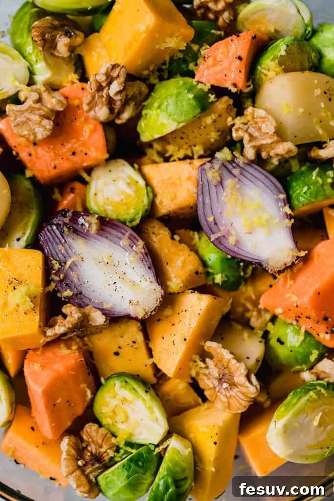 A large silver bowl filled with a mix of chopped raw vegetables including butternut squash, brussels sprouts, potatoes, and red onions, thoroughly tossed with olive oil, lemon zest, and chopped walnuts, ready for the oven.
