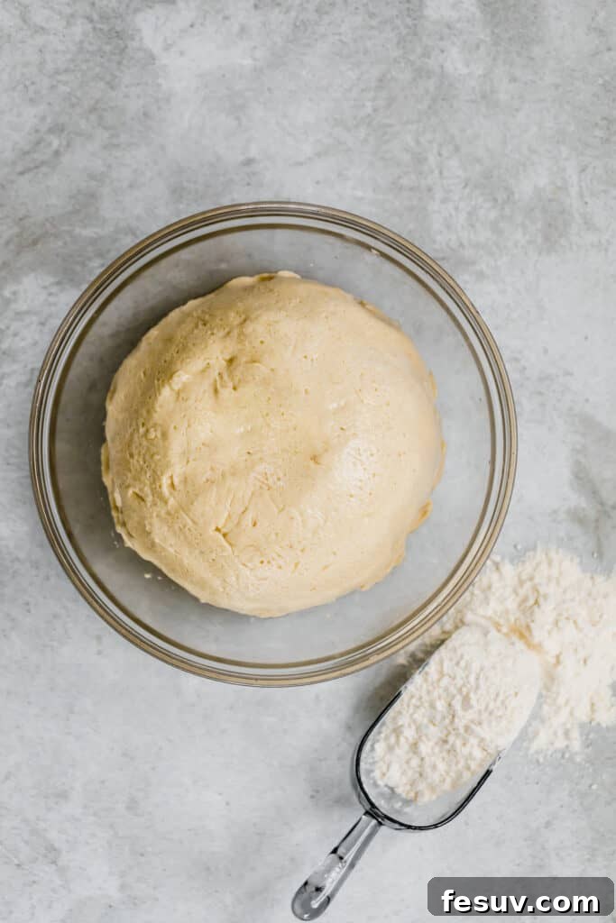 Smooth and elastic dinner roll dough resting in a clear glass bowl, with a flour scoop nearby