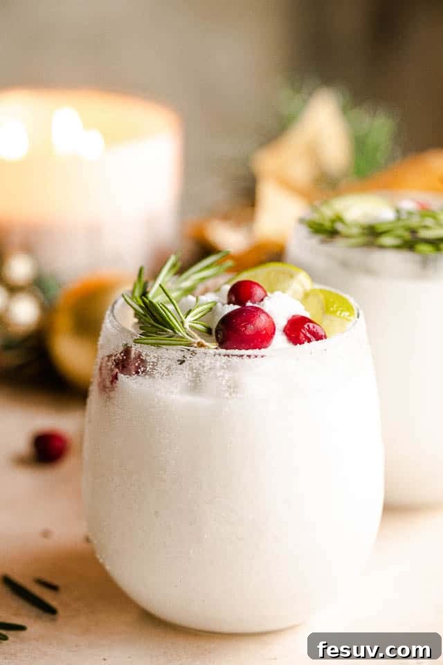 Drinking glass filled with frosty white margaritas, garnished with fresh lime slices, aromatic rosemary sprigs, and vibrant red cranberries, evoking a festive winter scene.