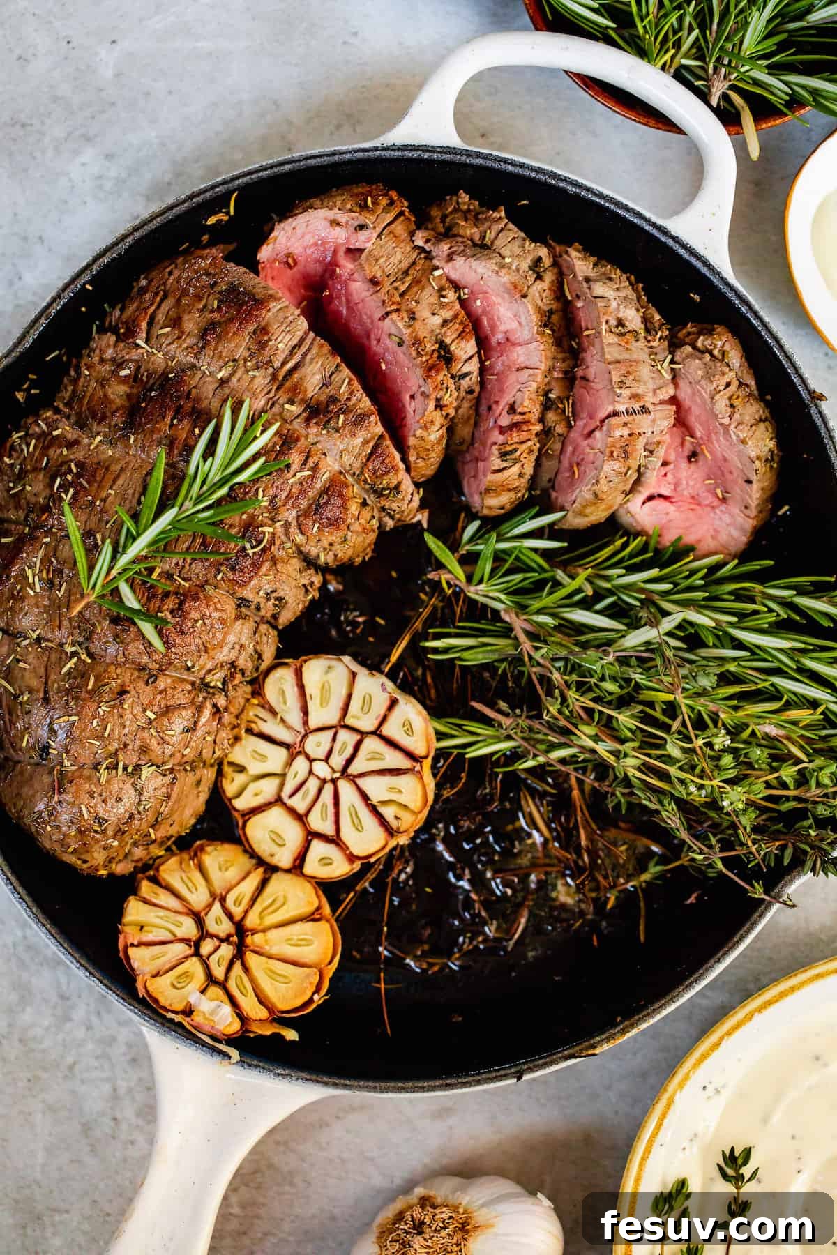A beautifully roasted beef tenderloin in a pan, garnished with whole garlic cloves and fresh sprigs of rosemary.