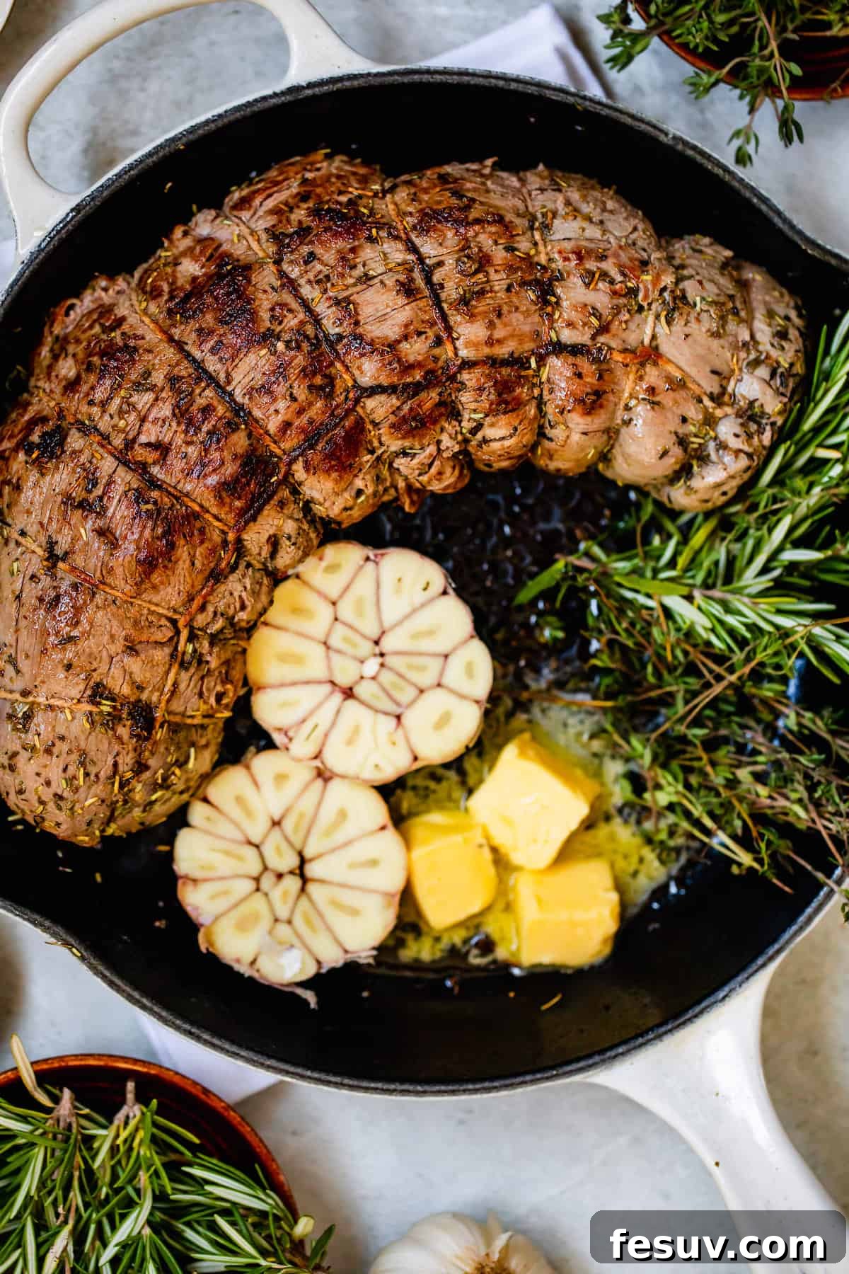 Raw beef tenderloin with garlic cloves, rosemary, and butter in a metal skillet, ready for searing.