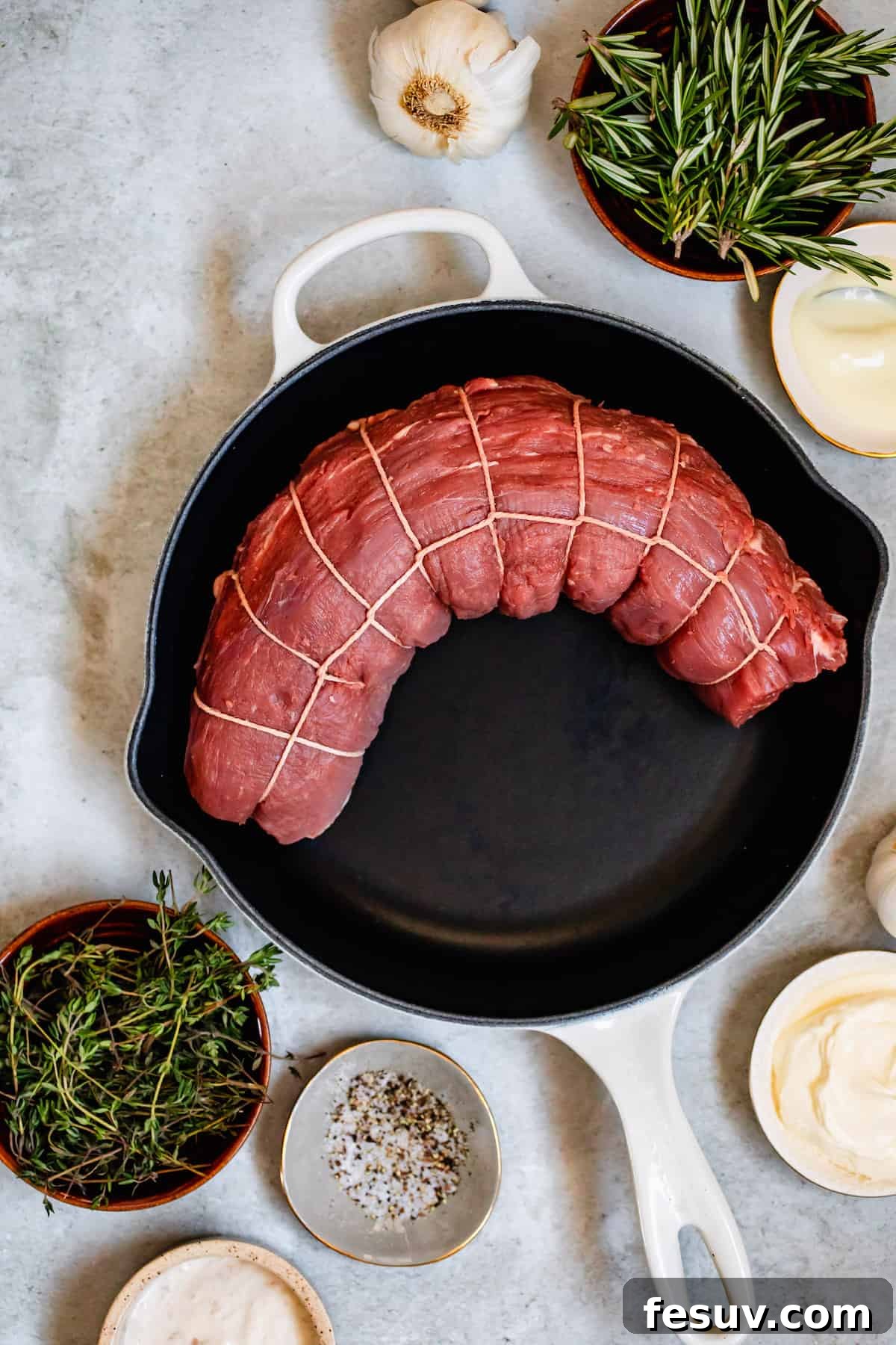 A tied beef tenderloin in a skillet, surrounded by all the recipe ingredients: dried herbs, garlic, salt, pepper, olive oil, and fresh sprigs of rosemary and thyme.