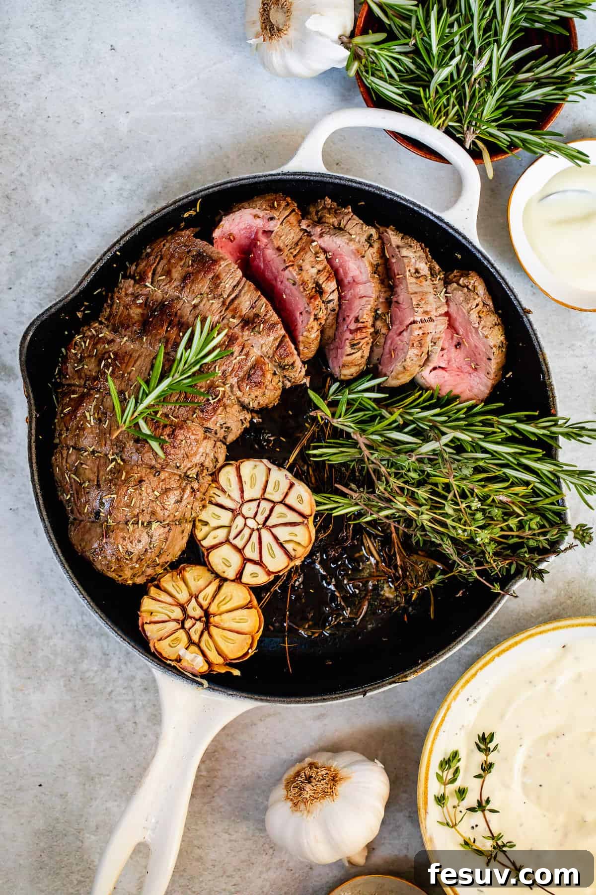 A white skillet containing the cooked beef tenderloin, garnished with garlic cloves and rosemary sprigs.