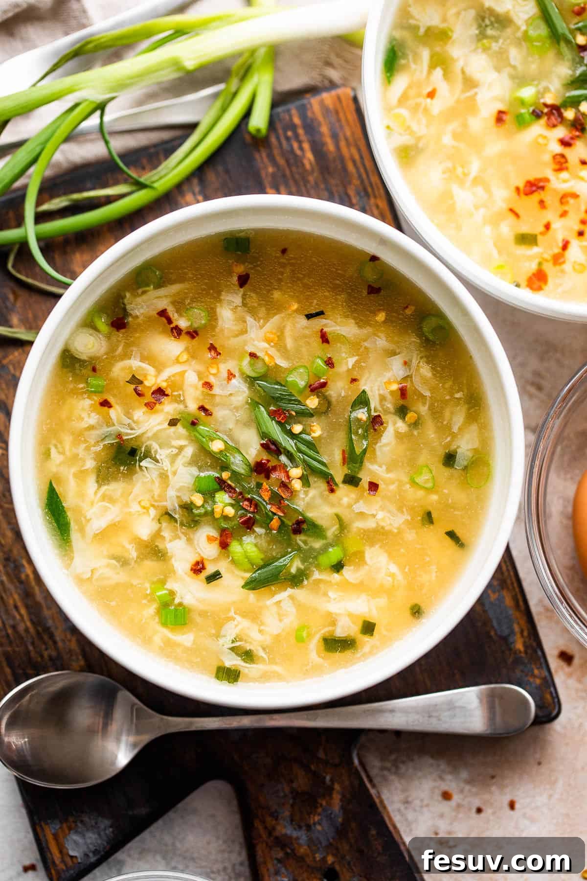 Simple and Quick Homemade Egg Drop Soup Recipe Two white bowls filled with golden egg drop soup, garnished with green onions and a hint of red pepper.