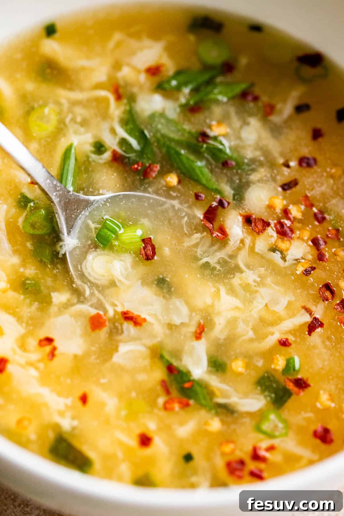 Garnished Egg Drop Soup Ready to Serve Close-up side view of a white bowl of egg drop soup, garnished with vibrant green onions and a sprinkle of red pepper flakes.