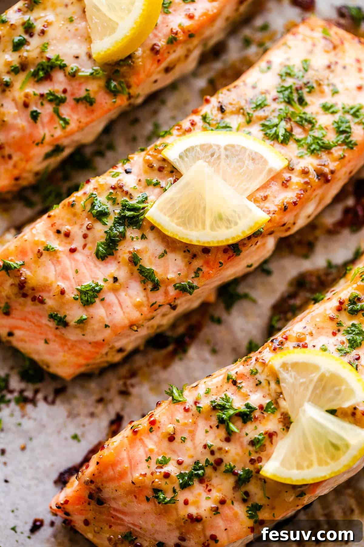 This baked Maple Mustard Salmon recipe is lightly seasoned, then brushed with a sweet and tangy sauce made from maple syrup and Dijon mustard! Close-up of baked maple mustard salmon fillets garnished with fresh parsley and bright lemon slices on a white plate.