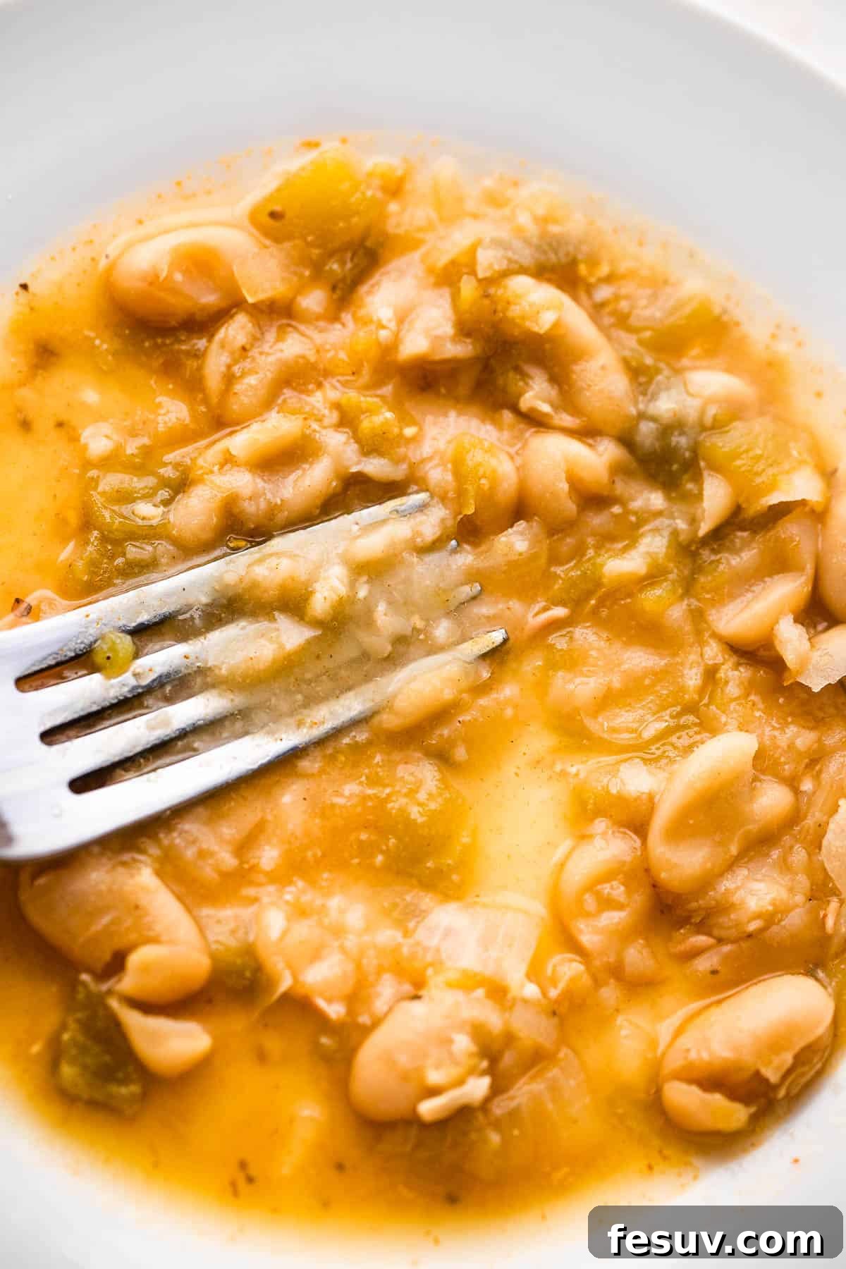 A close-up shot of a fork mashing cannellini beans on a white plate, demonstrating a technique for thickening chili.