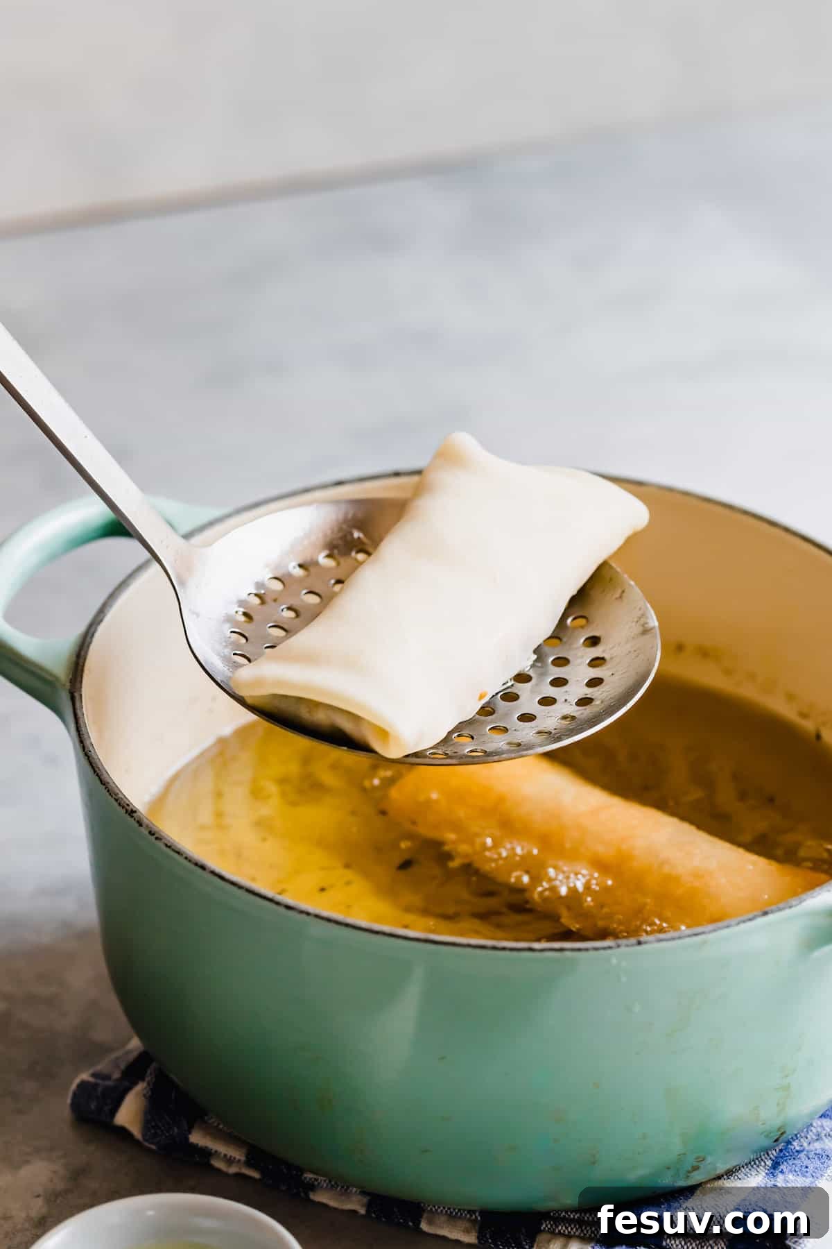 A Slotted Spoon Holding an Uncooked Egg Roll Over a Teal Frying Pan
