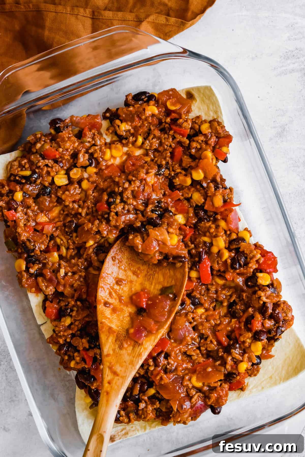Enchilada Lasagna 9 A wooden spoon delicately spreading an even layer of the savory Mexican lasagna filling over a bed of tortillas in a baking dish.