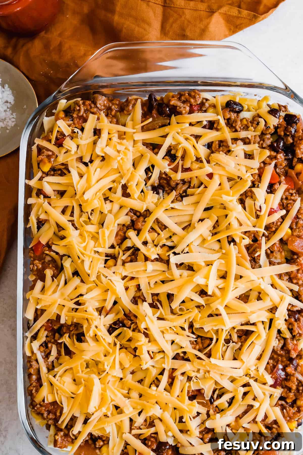 Enchilada Lasagna 10 A beautifully layered, unbaked Mexican Lasagna, showcasing its distinct layers of tortillas, meat filling, and a generous top layer of shredded cheddar cheese, ready for the oven.