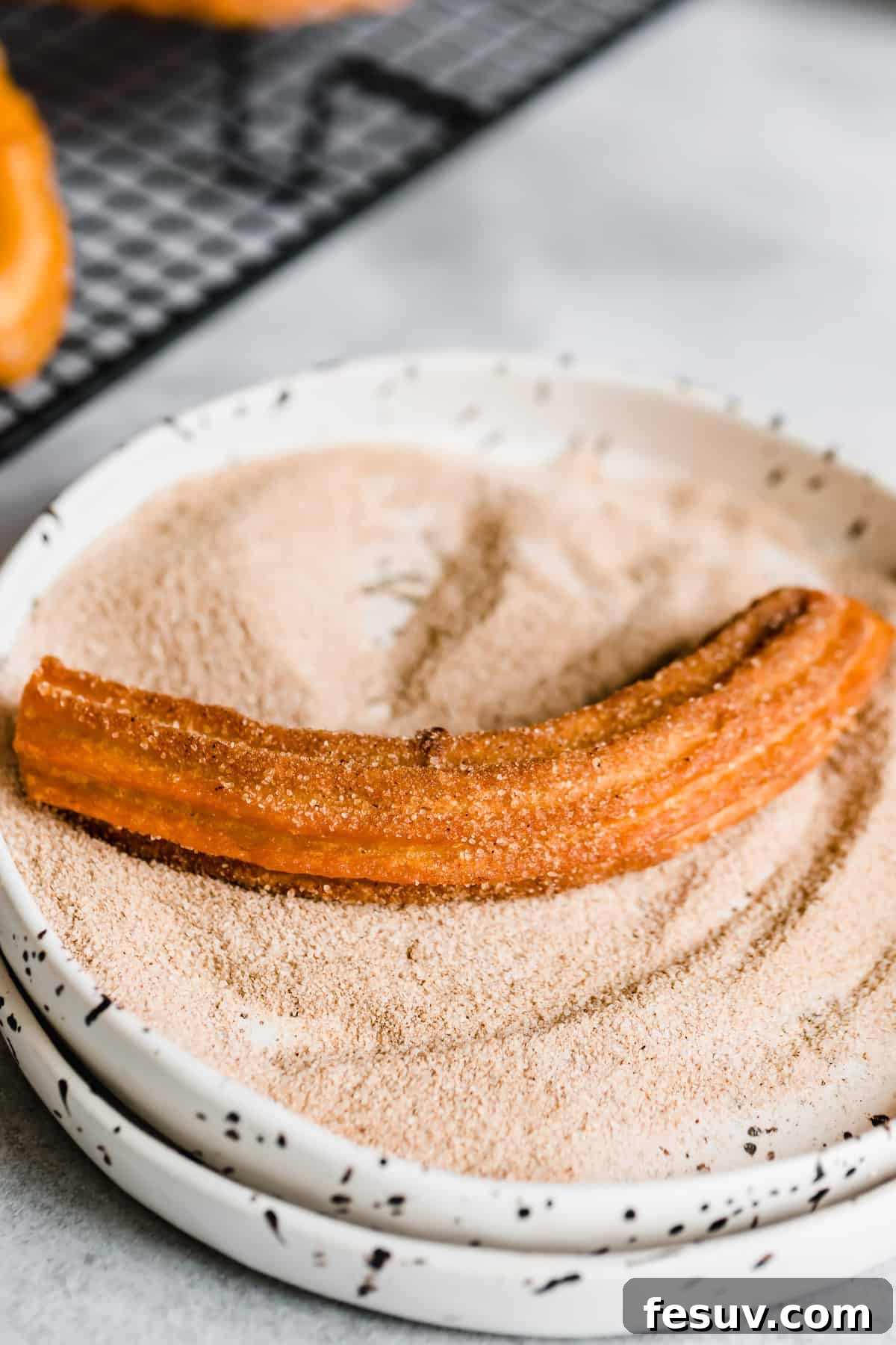 Perfect Homemade Churros 8 A single churro being rolled in a shallow bowl filled with cinnamon sugar for coating.