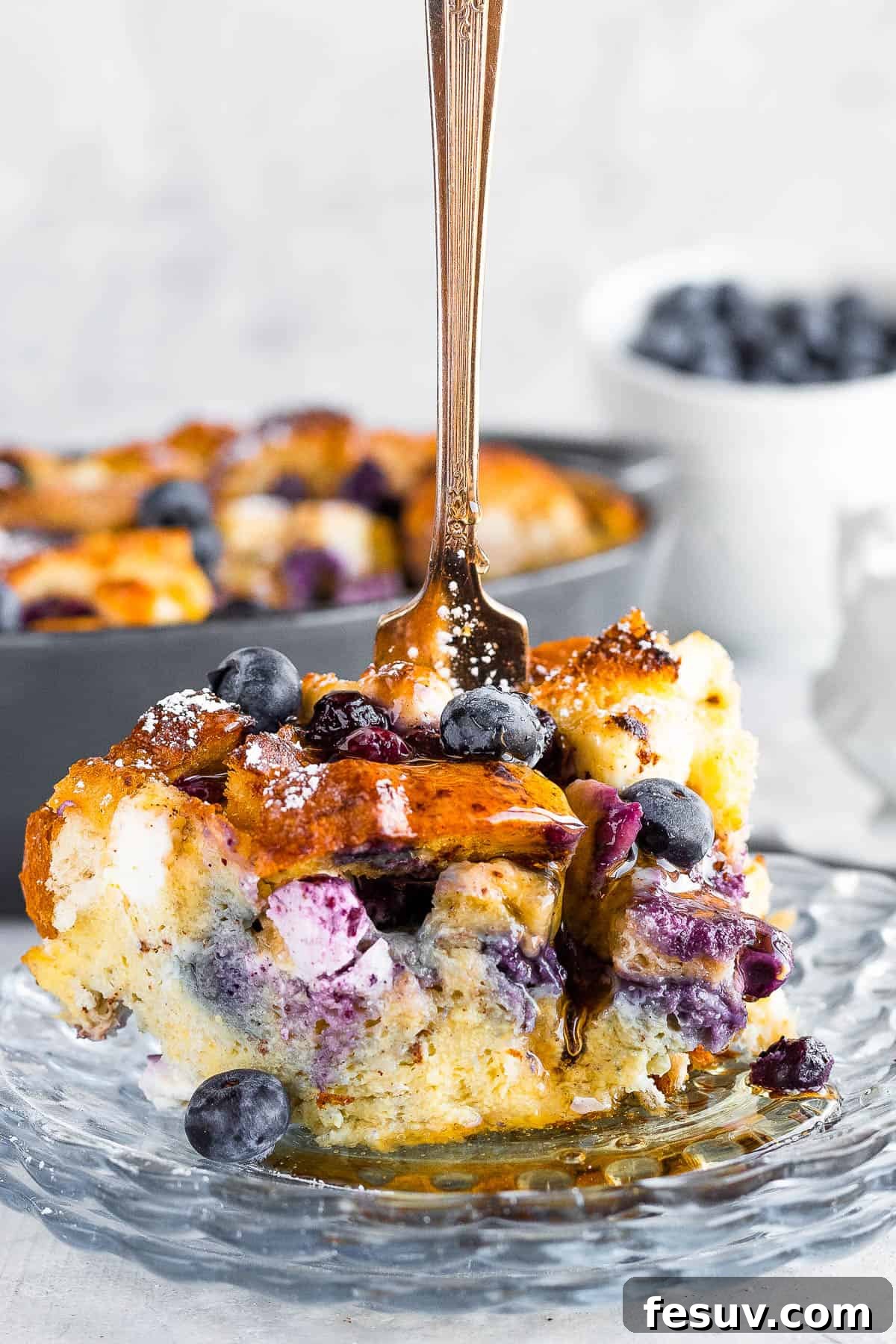Sweet Blueberry French Toast Bake 2 A Generous Piece of Blueberry French Toast Casserole with a Fork Sticking Upright Into it, Garnished with Powdered Sugar and Fresh Blueberries