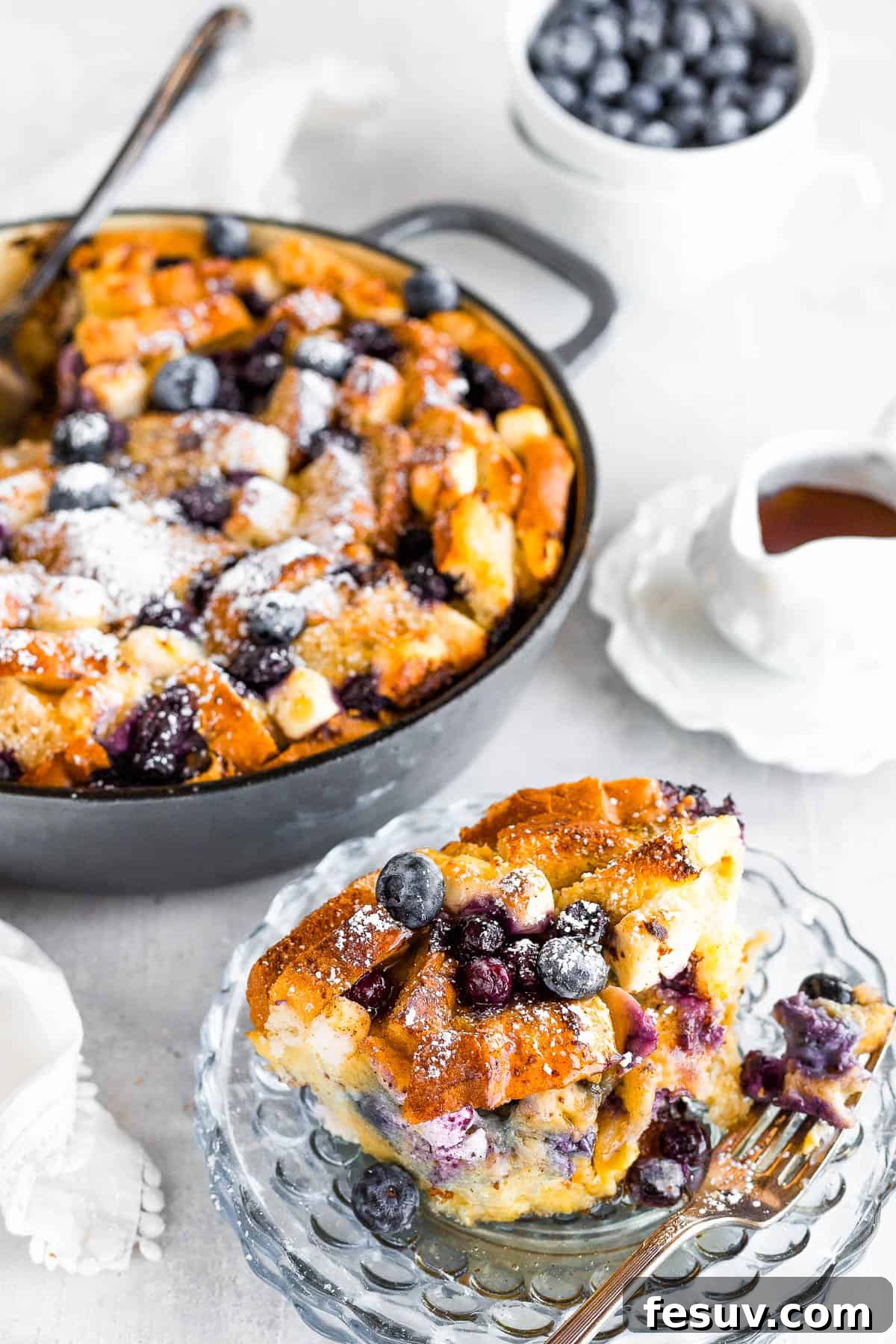 Sweet Blueberry French Toast Bake 3 A Serving of Baked Blueberry French Toast Bake on a Plate with the Full Casserole Behind it, Dusted with Powdered Sugar