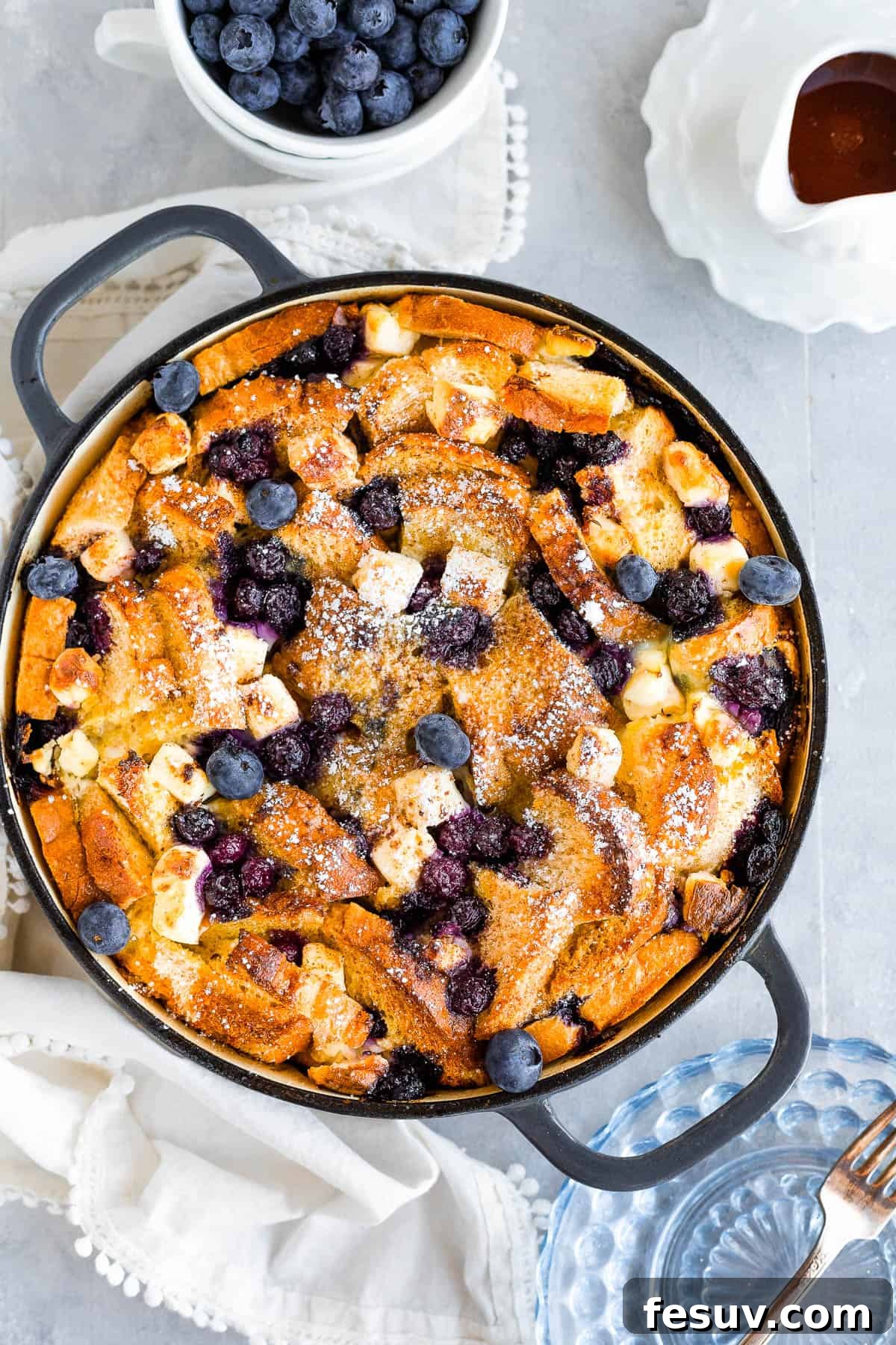 Sweet Blueberry French Toast Bake 9 A Bird's-Eye View of a Delicious Baked Blueberry French Toast Casserole Beside a Small Cup of Fresh Blueberries and a Spouted Measuring Cup Filled with Maple Syrup