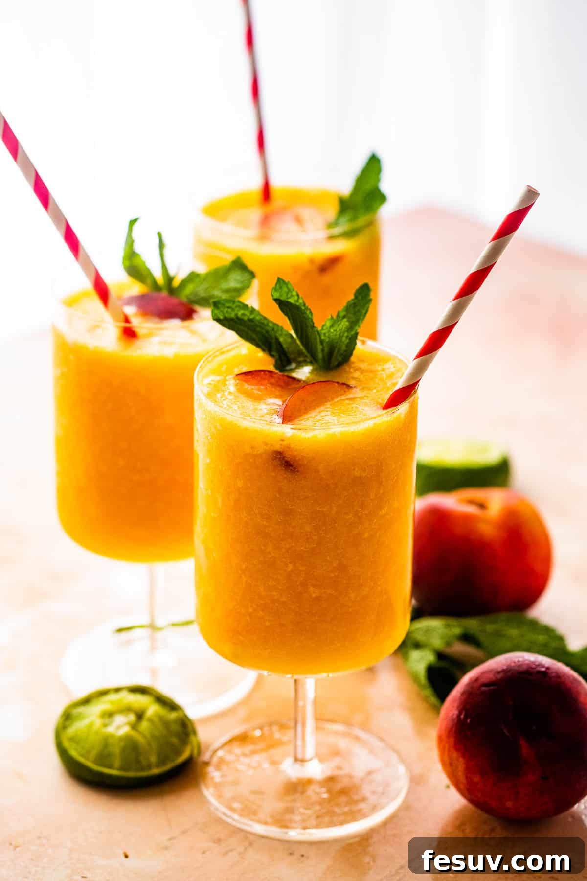 three glasses filled with peach daiquiris and garnished with mint, peach slices, and straws