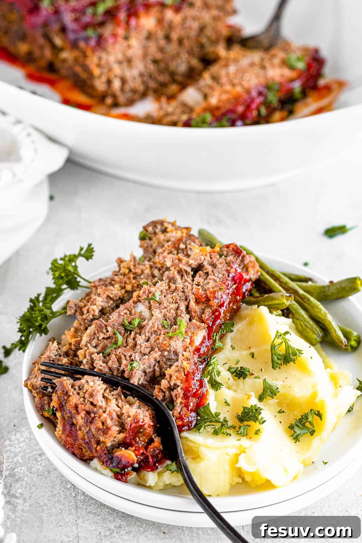 A family-friendly classic, this easy meatloaf recipe gets extra flavor from barbecue sauce, plenty of garlic, and Italian seasoning. A fork digging into a thick slice of perfectly baked meatloaf, served on a plate with mashed potatoes and green vegetables.