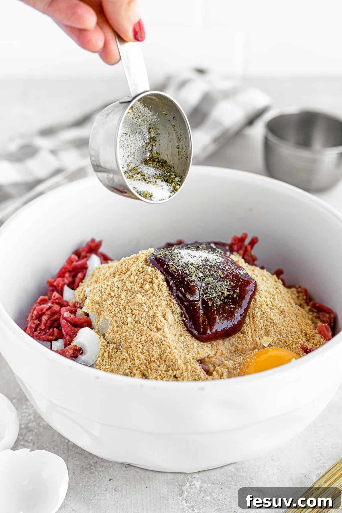 A family-friendly classic, this easy meatloaf recipe gets extra flavor from barbecue sauce, plenty of garlic, and Italian seasoning. A large bowl containing the meatloaf mixture, ready to be shaped, next to a glass loaf pan on a gray countertop.