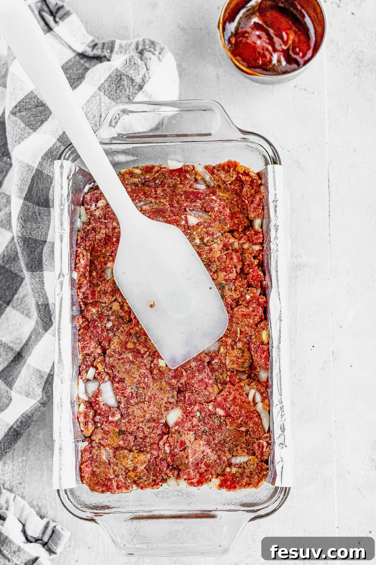 A family-friendly classic, this easy meatloaf recipe gets extra flavor from barbecue sauce, plenty of garlic, and Italian seasoning. A spatula gently spreading the prepared meatloaf mixture evenly into a foil-lined loaf pan.