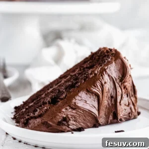 A piece of frosted chocolate cake on a plate with the rest of the cake behind it