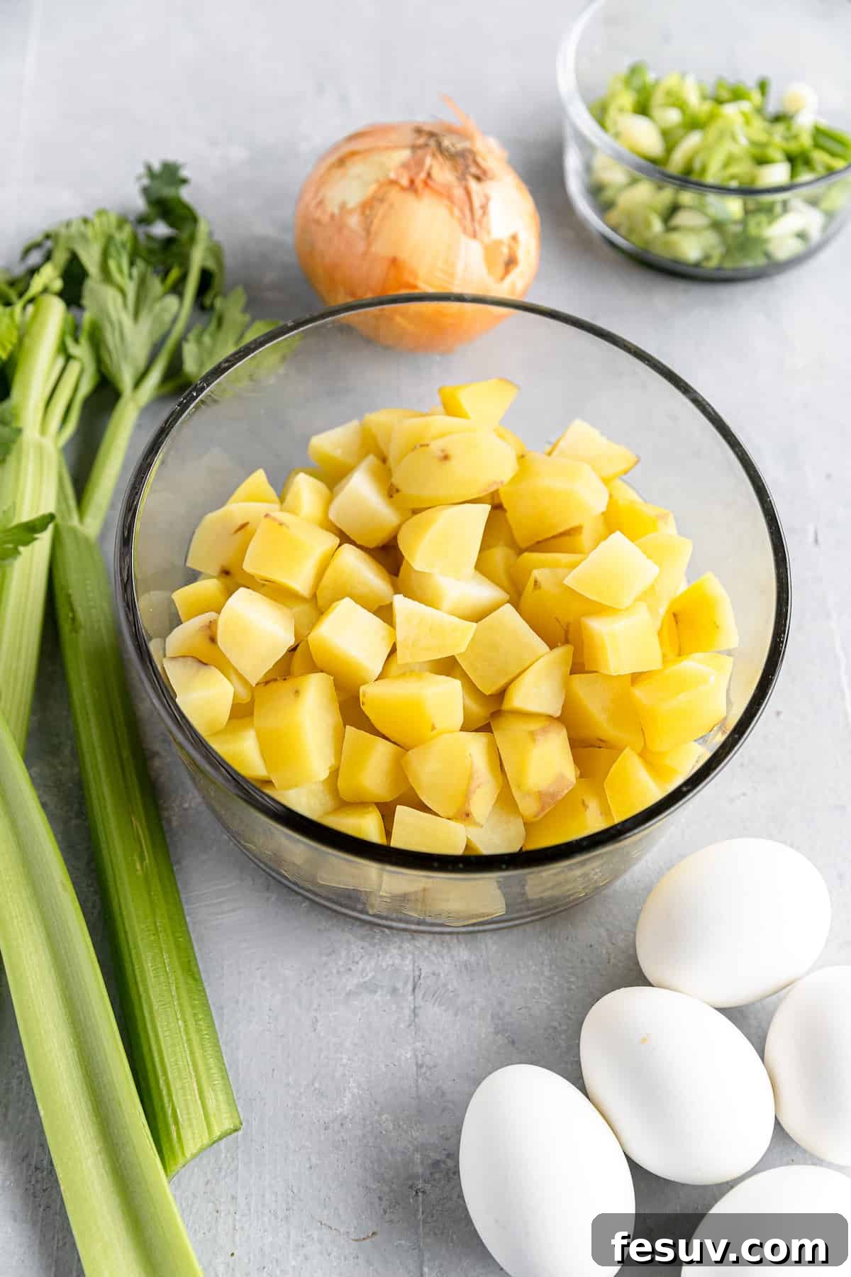 America's Favorite Potato Salad 4 A Vibrant Arrangement of Fresh Potato Salad Ingredients on a Gray Countertop, Featuring Diced Potatoes, Hard-Boiled Eggs, and a Whole Yellow Onion, Ready for Preparation.