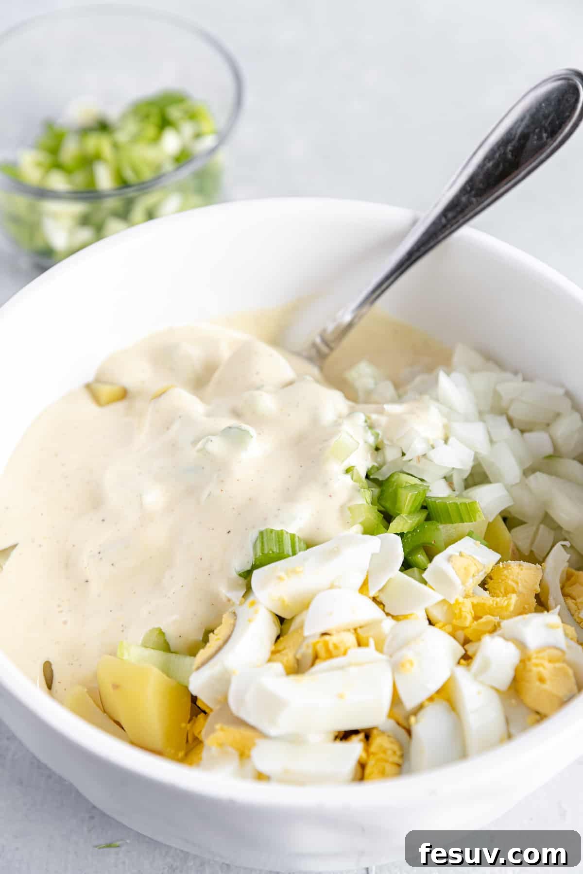 America's Favorite Potato Salad 6 The Creamy Mayonnaise-Based Dressing Being Poured Over the Mixture of Diced Potatoes, Celery, Onion, and Hard-Boiled Eggs in a White Bowl, Ready to Be Combined into a Classic Potato Salad.