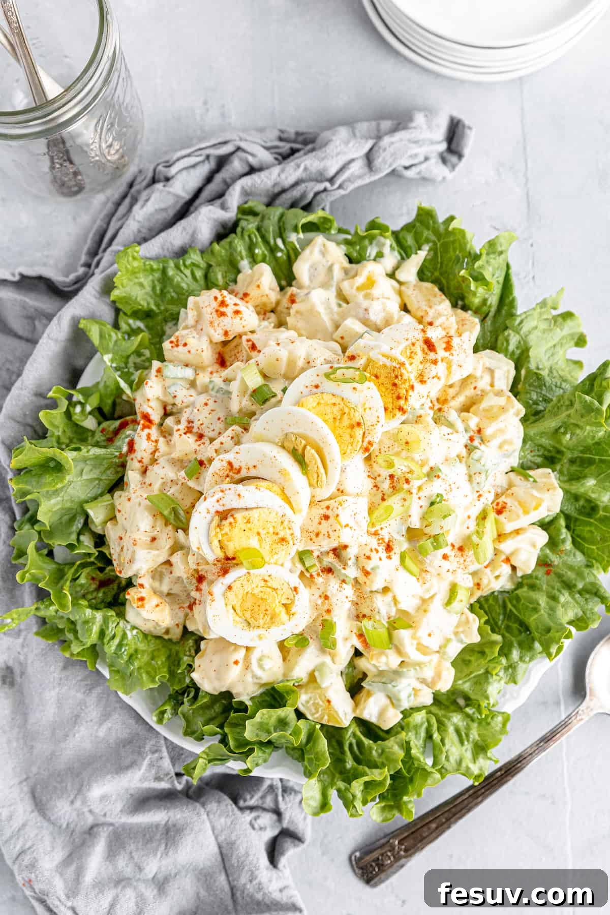 America's Favorite Potato Salad 9 An Inviting Overhead View of a Generous Portion of Creamy American Classic Potato Salad, Elegantly Arranged on a Lettuce-Lined Plate, Resting Beside a Stylish Gray Kitchen Towel.