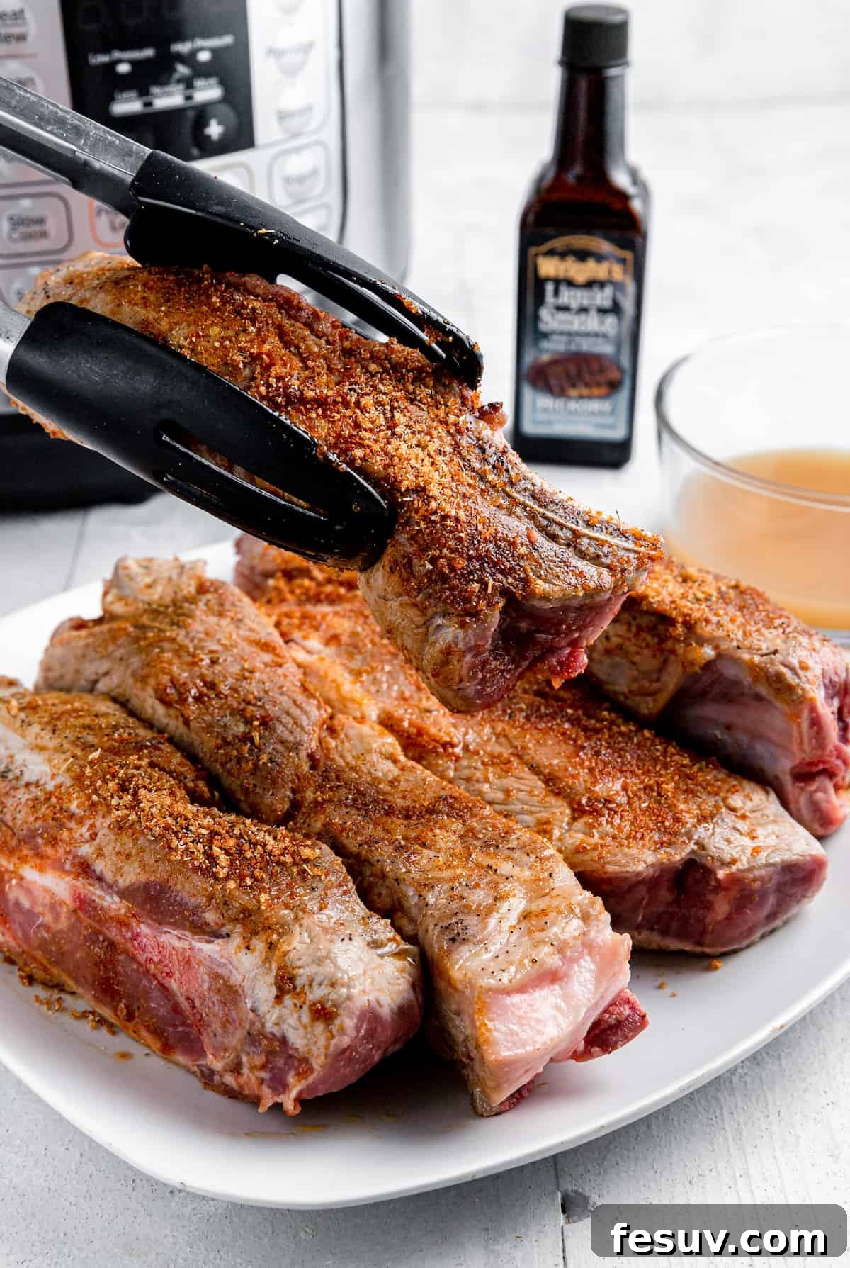 Fall-Off-The-Bone Instant Pot Ribs 5 A pair of tongs holding a generously seasoned pork rib over a plate full of other seasoned ribs, ready for cooking