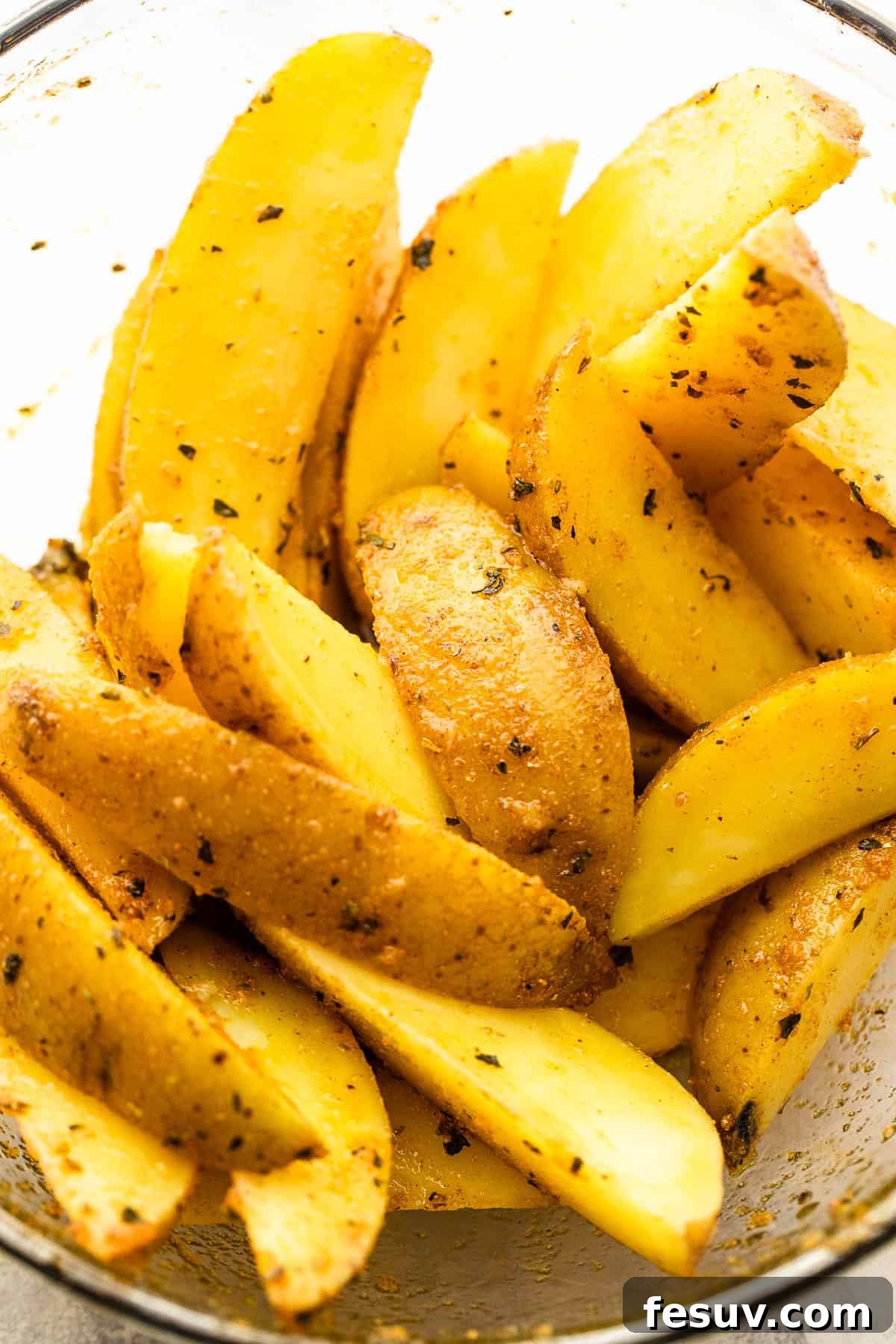 Preparing potato wedges with a flavorful herb and olive oil coating. Cut Russet potatoes in a glass bowl, generously seasoned with herbs and olive oil.