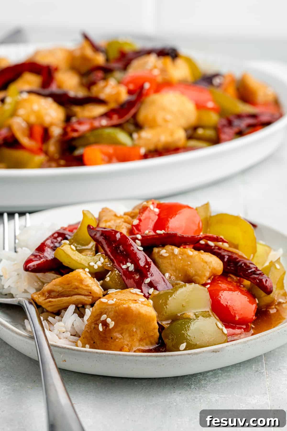 Two hands stirring Szechuan Chicken in a large frying pan, showing cooked chicken pieces, colorful bell peppers, and onions coated in a rich, dark sauce.