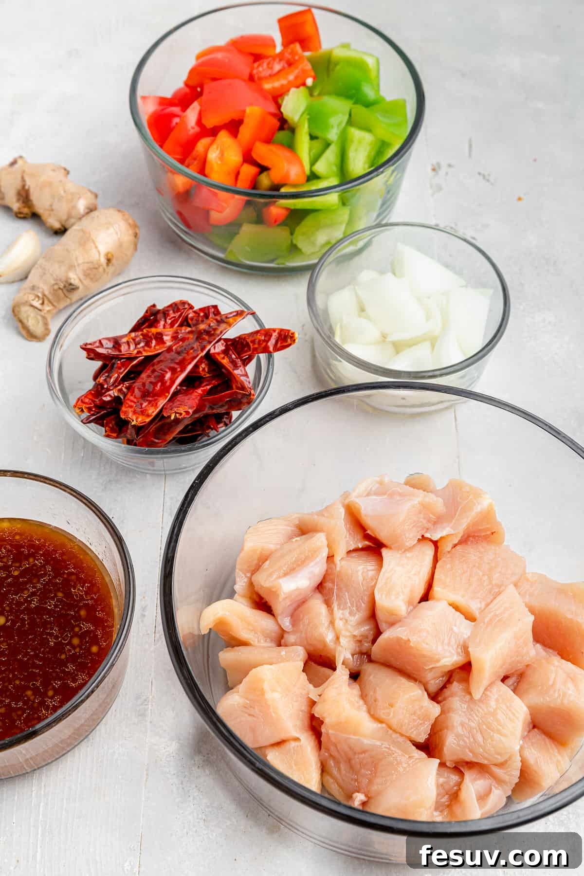 A visual guide to Szechuan Chicken ingredients, arranged clockwise: chopped bell pepper, chopped onion, cubed chicken, Szechuan sauce in a bowl, dried chilies, fresh ginger, and minced garlic.