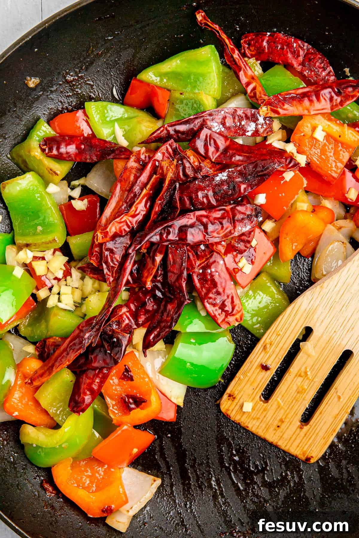 Chopped bell peppers, onions, minced garlic, ginger, and small dried chilies are sizzling in a hot pan during the stir-fry process.