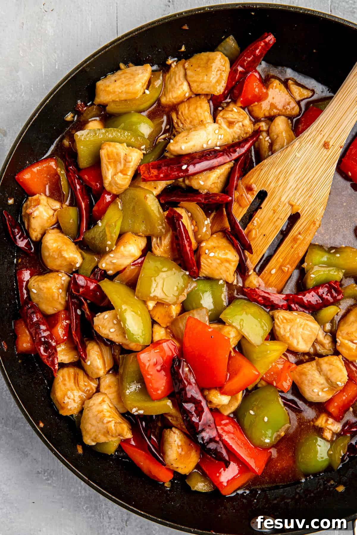 Szechuan chicken stir-fry served in a cast-iron skillet, with a wooden spatula poised to serve. The dish is rich with chicken, bell peppers, and chilies.