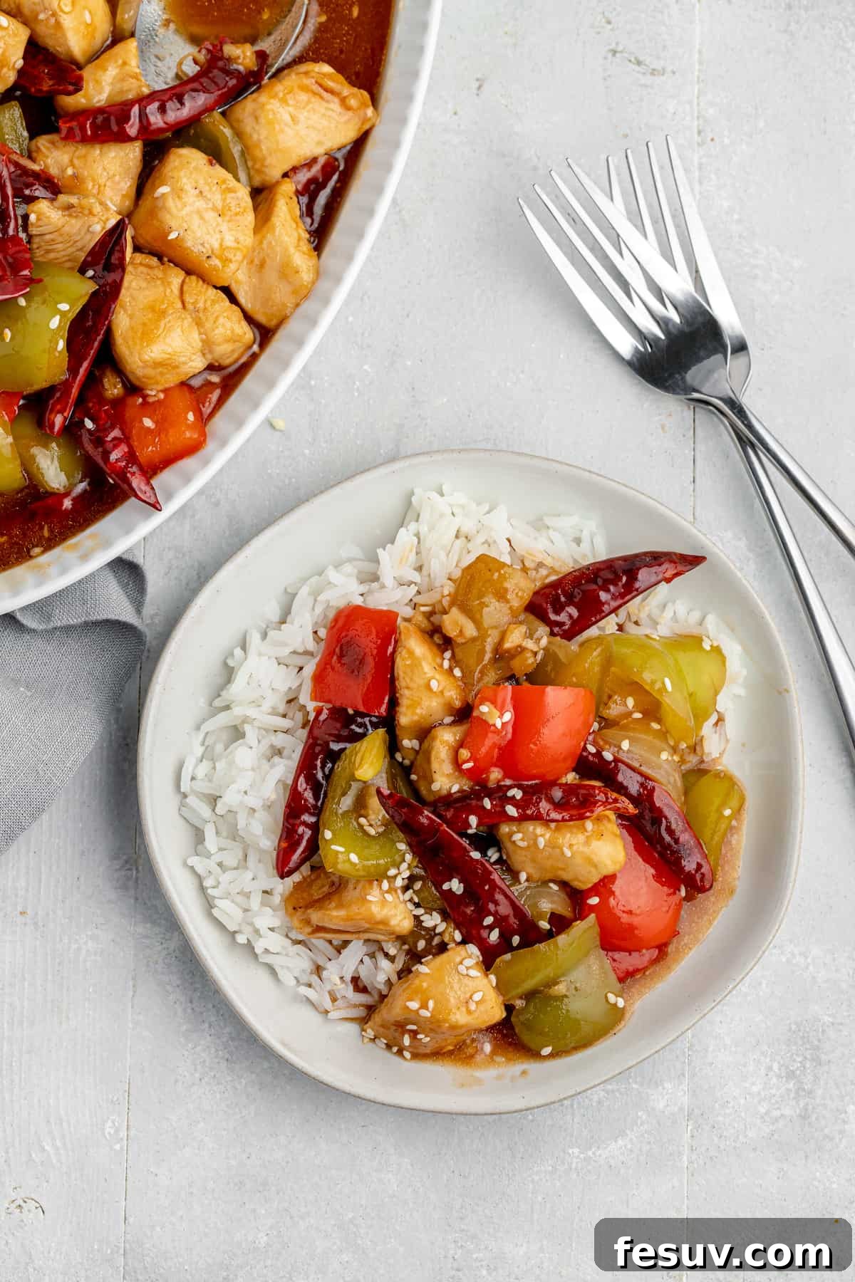 A beautifully plated serving of Szechuan chicken stir-fry over white rice, with a large serving platter and two sets of chopsticks on the table, invitingly set for a meal.