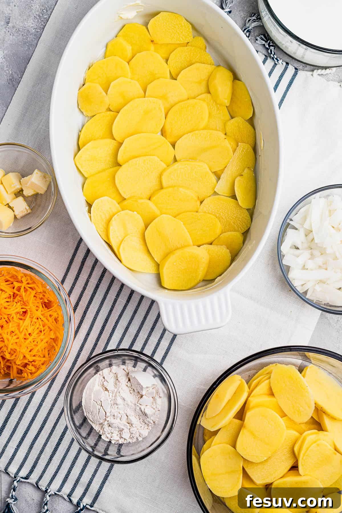 Tender sliced potatoes layered with cheddar cheese and diced onions to make a scalloped potatoes casserole. Sliced potatoes layered in a baking dish, ready for the next steps.