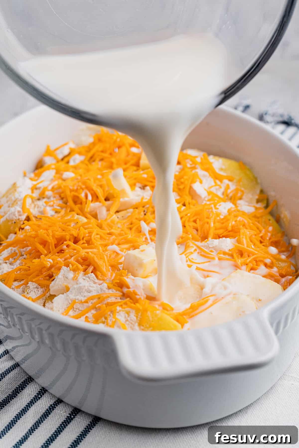 Tender sliced potatoes layered with cheddar cheese and diced onions to make a scalloped potatoes casserole. Pouring milk over sliced potatoes and shredded cheese in a baking dish, preparing for baking.